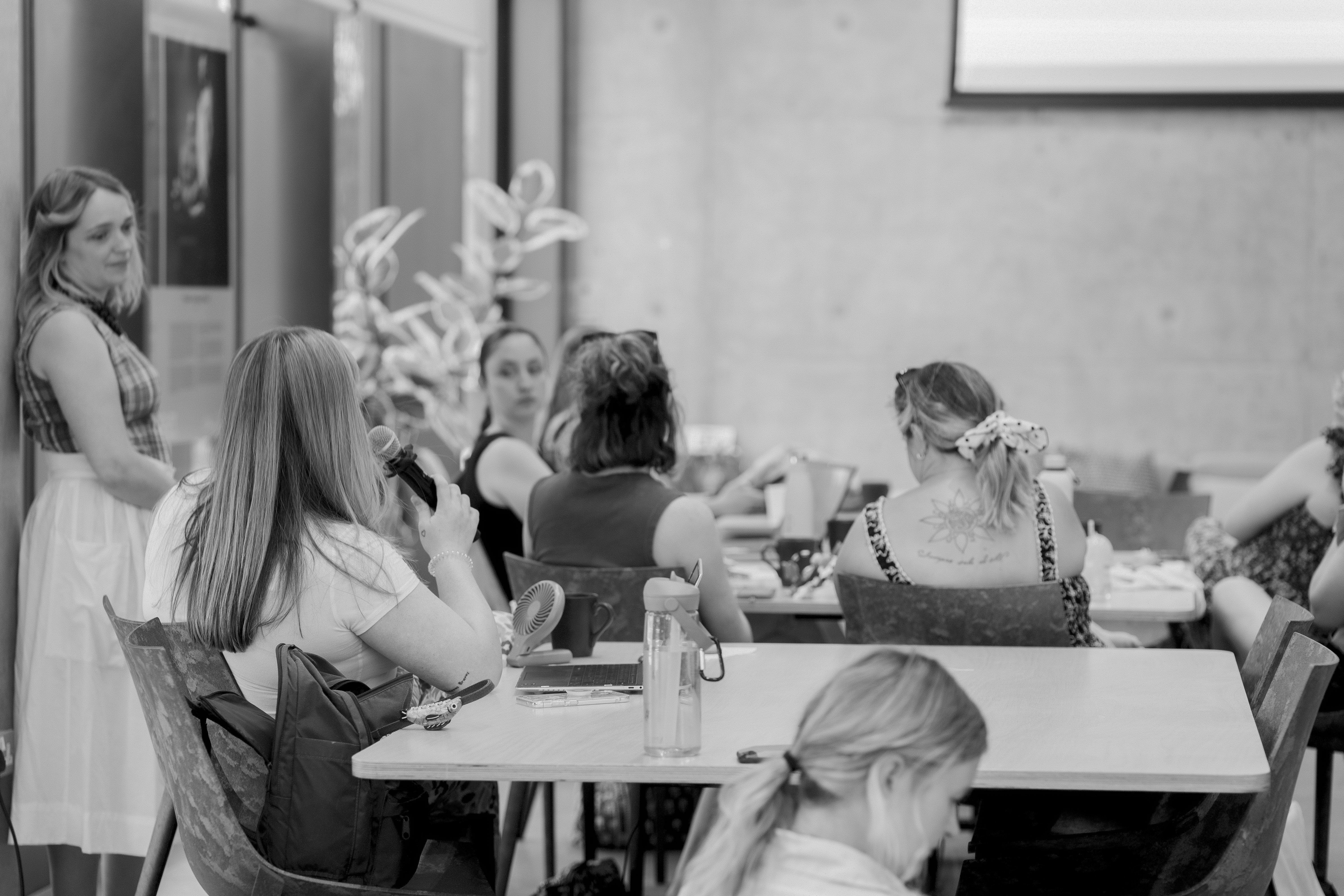 Women at tables during an AI on Maternity Leave event at Plus X Innovation in Brighton, one holding a microphone