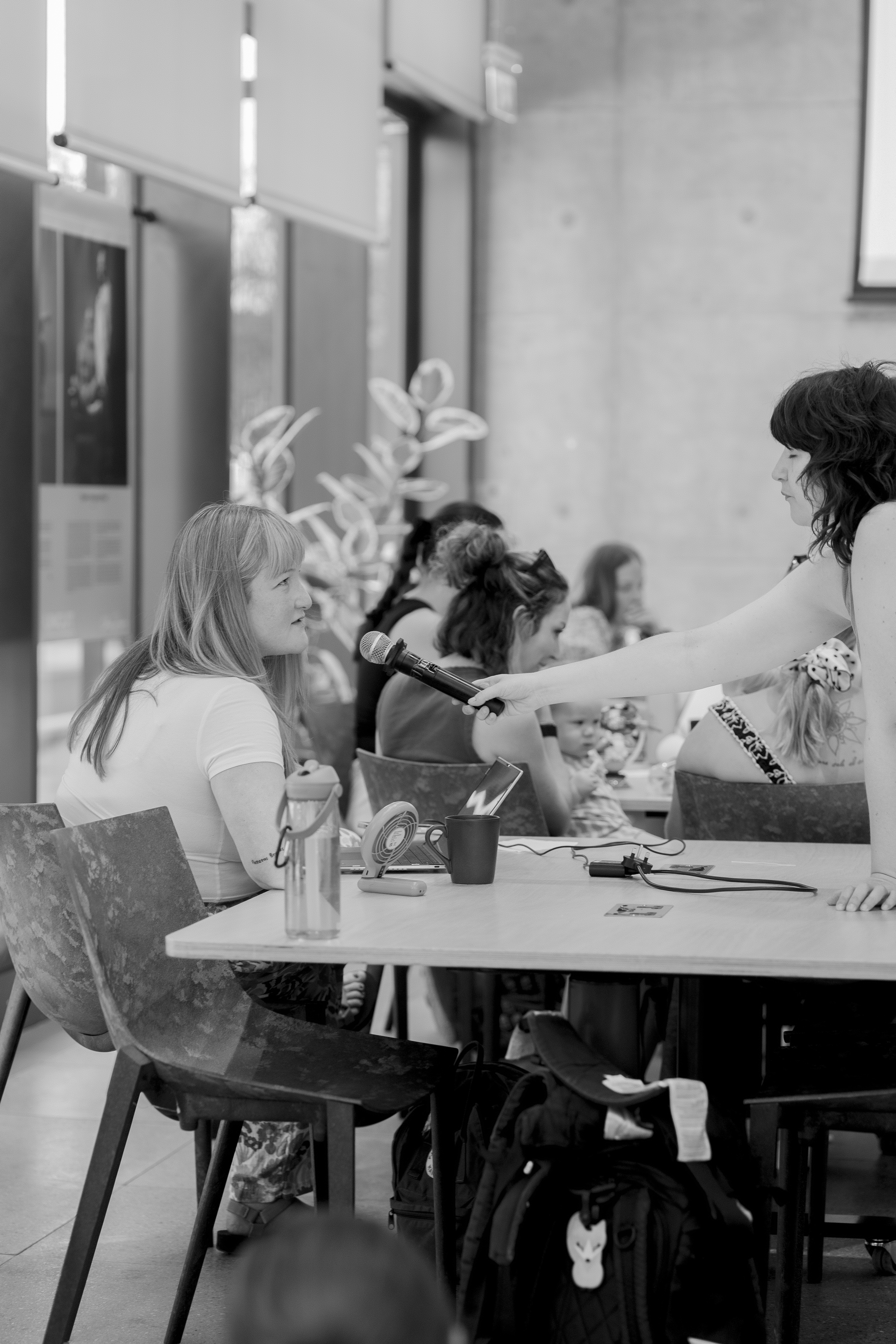 A woman being handed a microphone at an AI on Maternity Leave event, with other mums and a baby at the table behind her