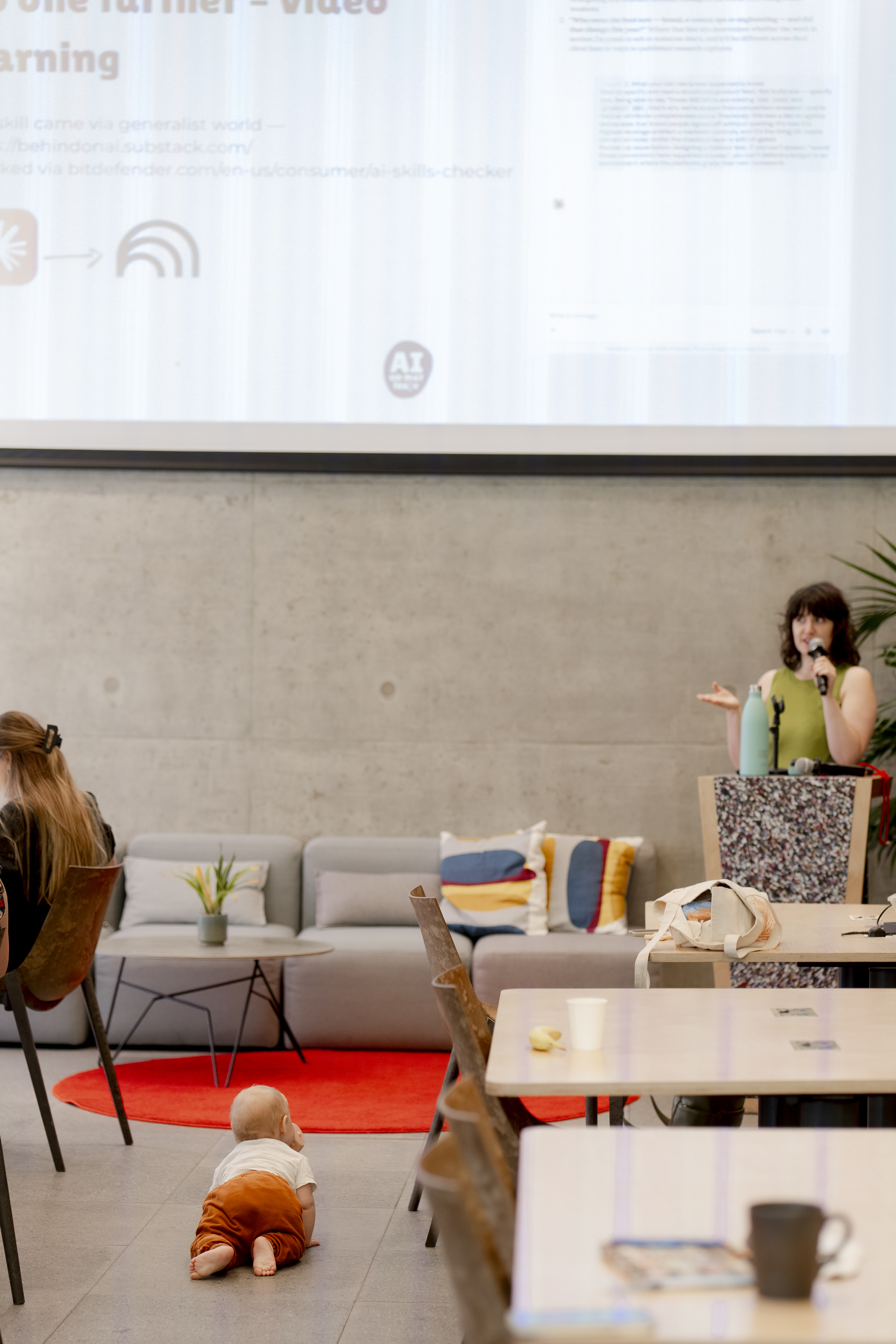A speaker presenting at an AI on Maternity Leave event while a baby crawls across the floor