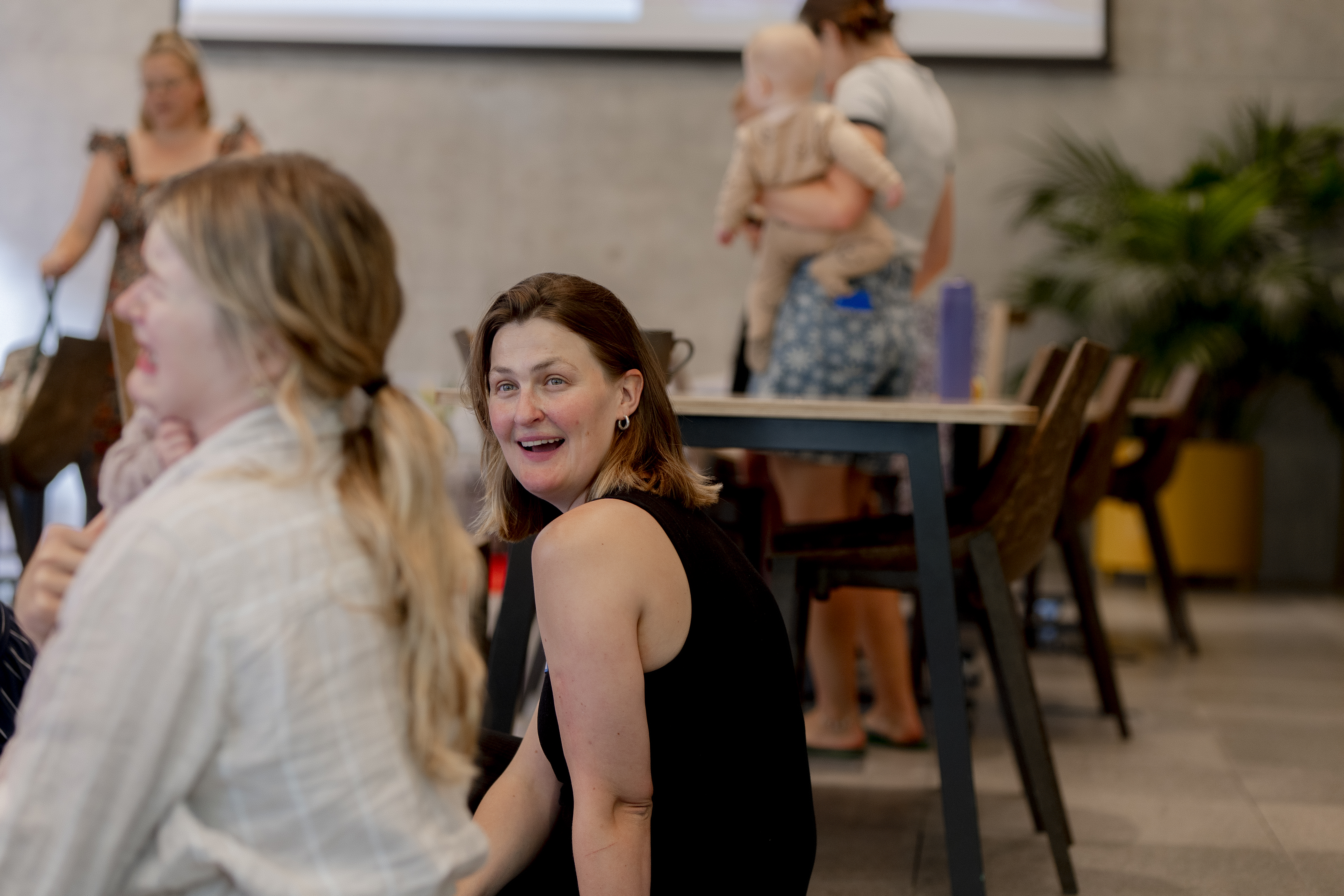 A woman turning to smile at the camera while chatting with others at an AI on Maternity Leave event, a baby being carried in the background