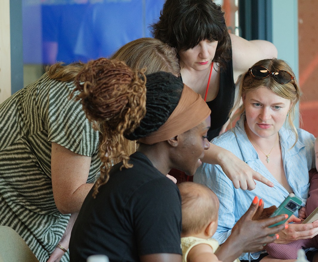 Four women huddled around a phone, one with a baby on her lap, at an AI on Maternity Leave event