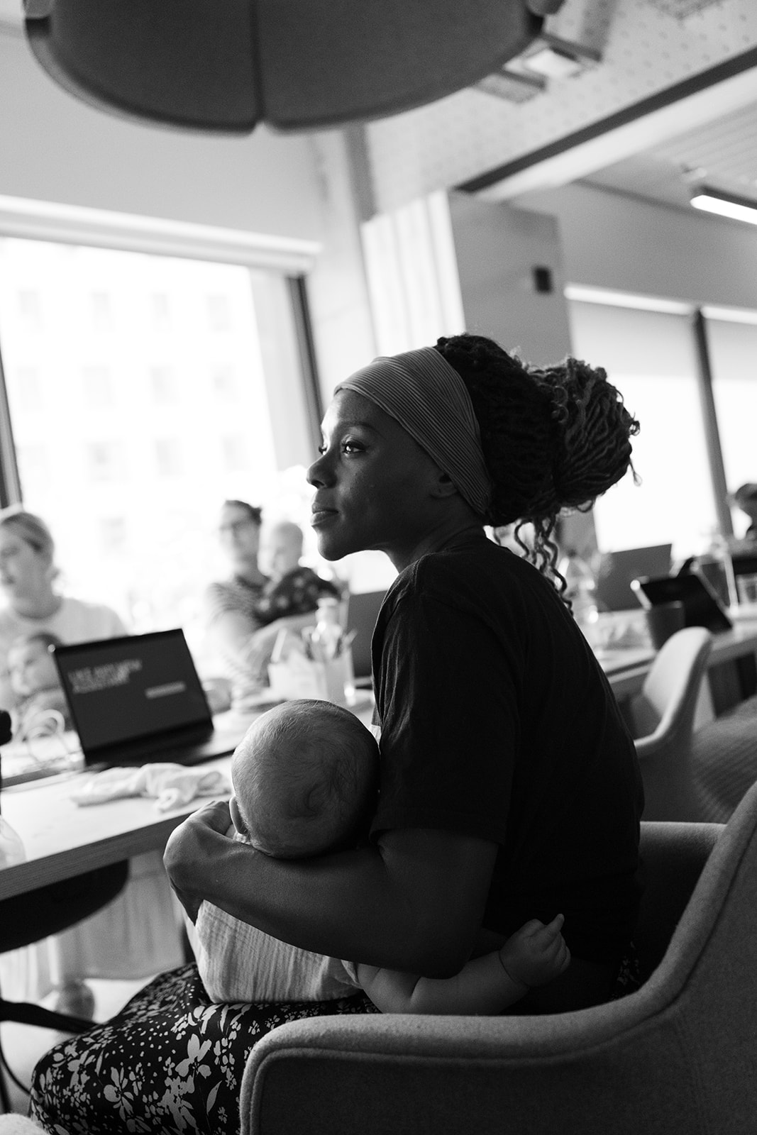 A woman in a headwrap holding her sleeping baby, listening at an AI on Maternity Leave event