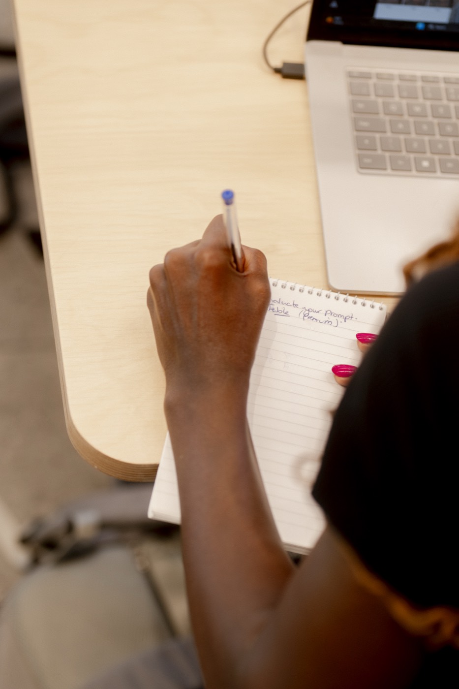 A woman taking handwritten notes beside her laptop, planning her prompts